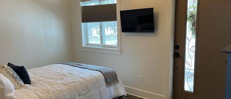 Un dormitorio con cama, televisor montado en la pared, ventana con persianas y puerta con panel de vidrio.