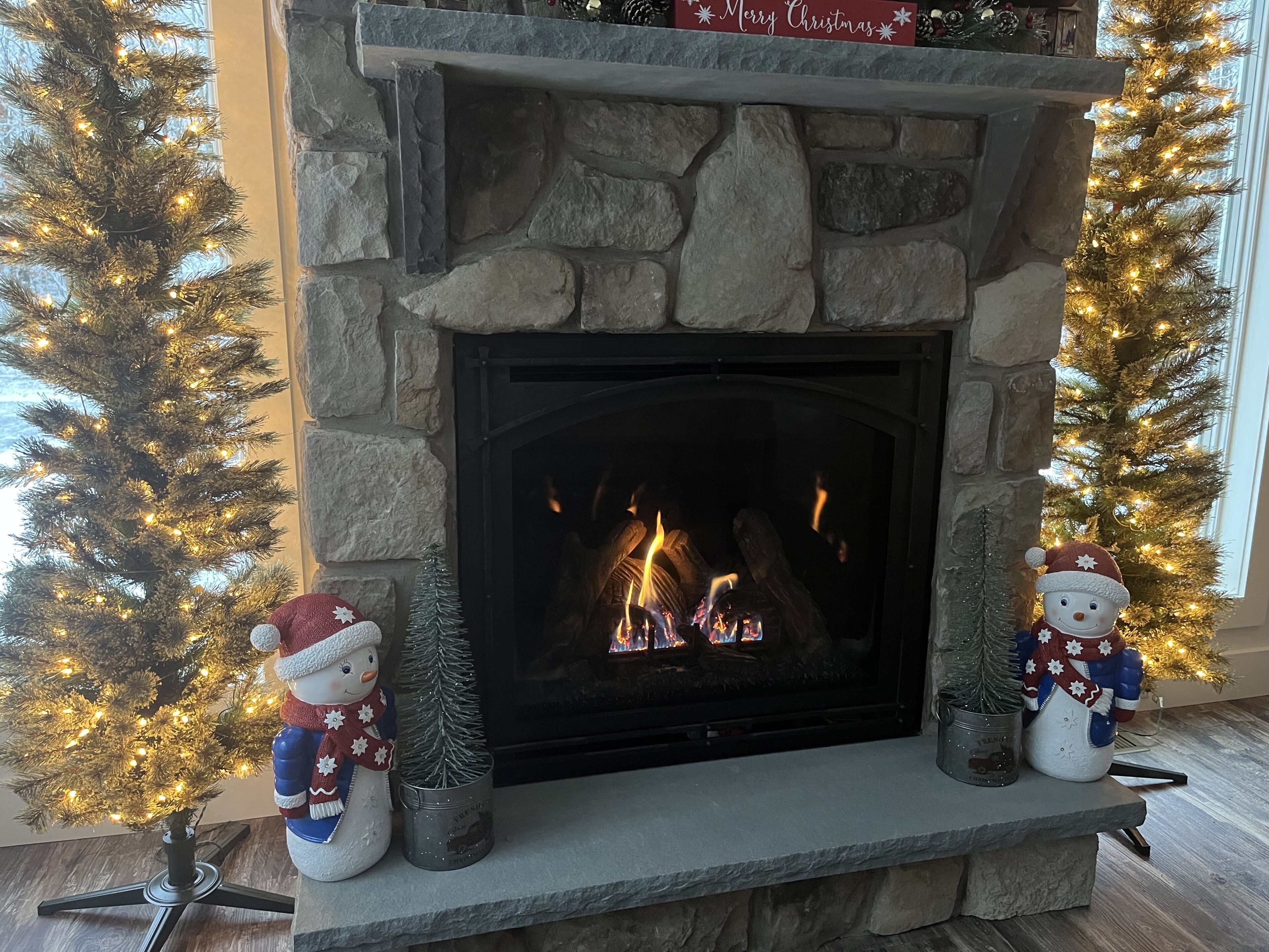 A stone fireplace with a lit fire, flanked by two snowman decorations and a Christmas tree with lights.