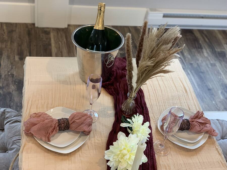 Una tavola apparecchiata con piatti bianchi, tovaglioli rosa, una bottiglia di champagne e un vaso con pampas e fiori bianchi.