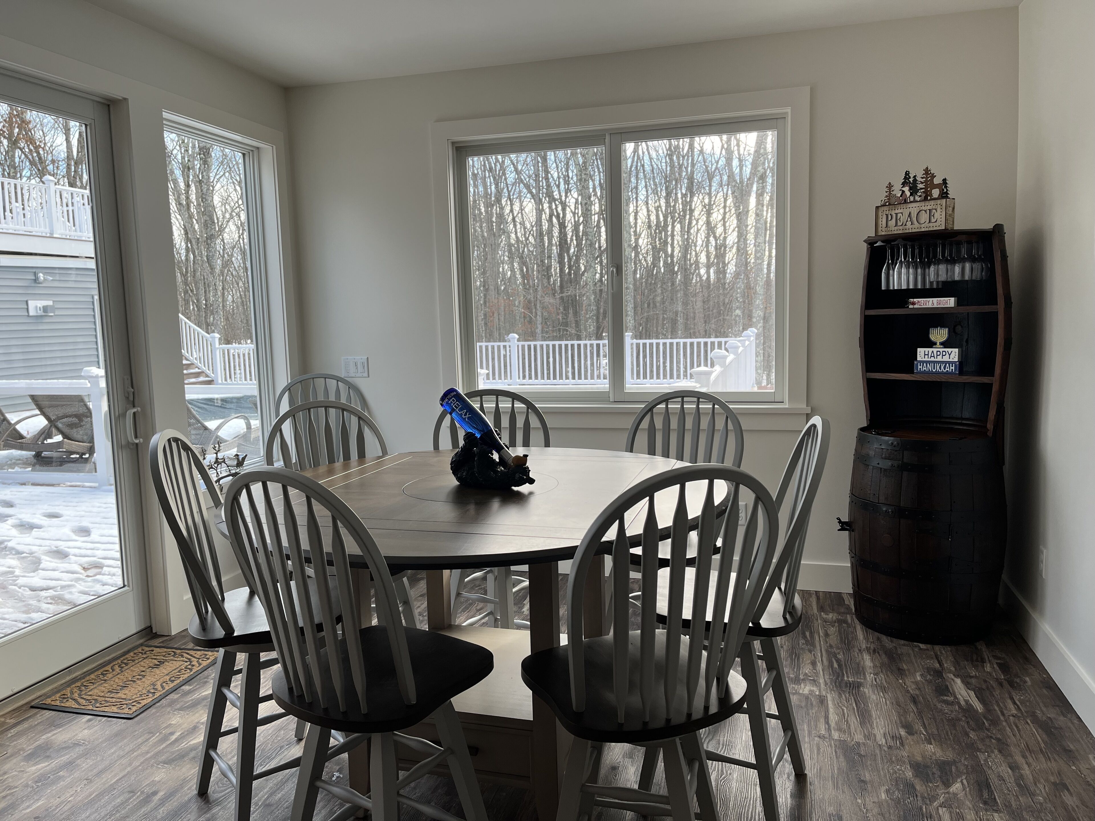 A dining area with a table and chairs, a large window with a view of a deck, and a shelf with holiday decorations.