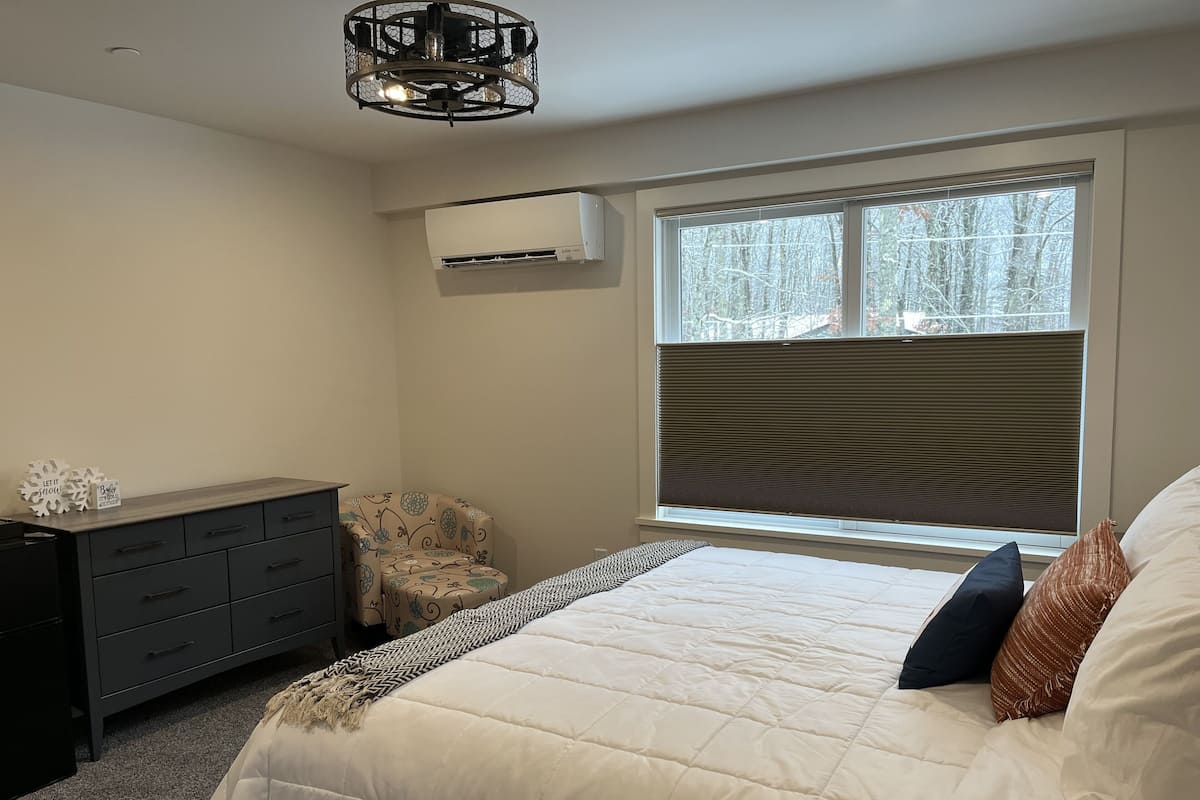 Une chambre à coucher comprenant un lit, une commode, un climatiseur, une fenêtre avec des stores et un luminaire au plafond.
