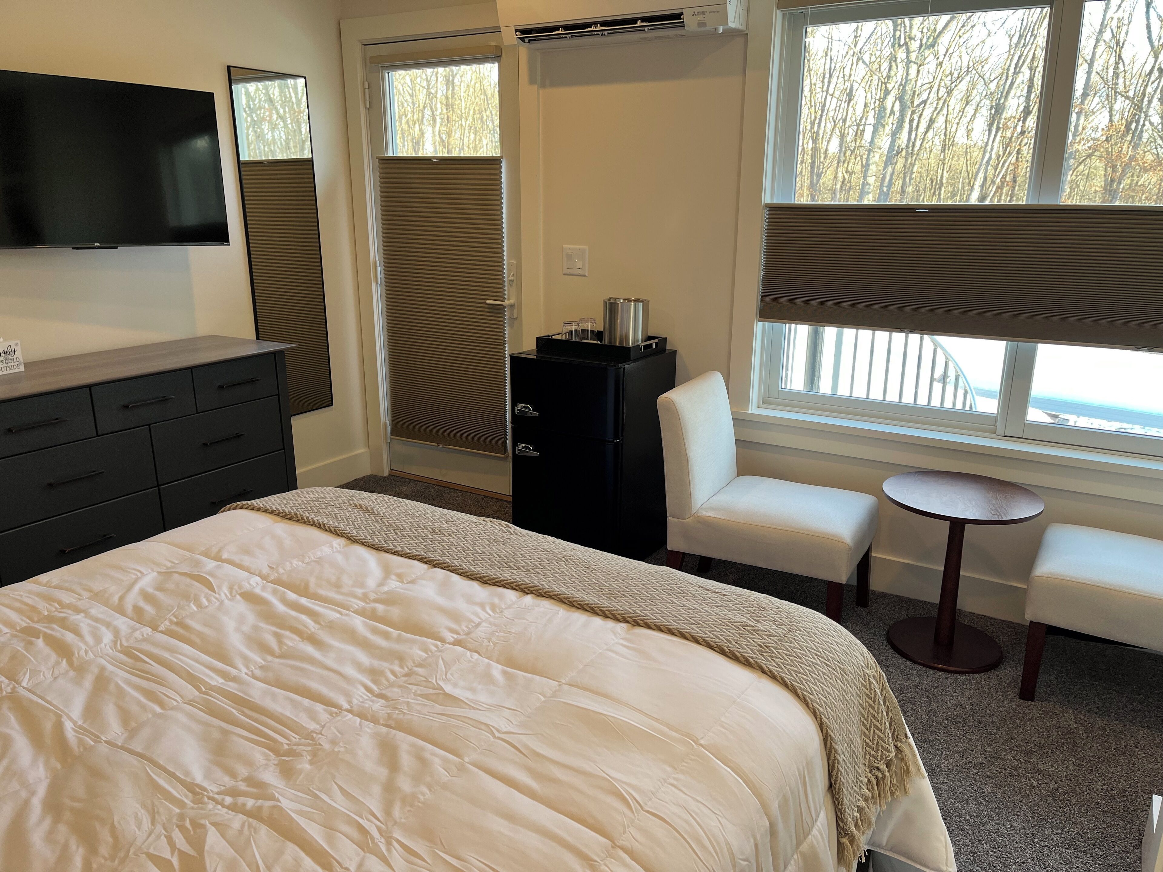 A hotel room with a large bed, a TV, a desk, a chair, a small table, and a window with blinds.