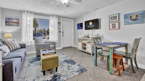 Un salon comprenant un canapé, un pouf et un coin repas avec une table et des chaises. On y trouve également un ventilateur de plafond, un téléviseur et des œuvres d’art accrochées aux murs.