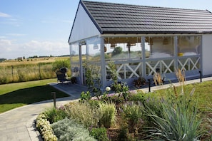 A covered outdoor seating area with a grill and a chair, surrounded by a garden with various plants and flowers.
