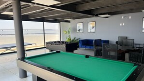 Een pooltafel in een moderne ruimte met een blauwe bank en stoelen, grote ramen en uitzicht op het strand.