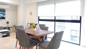 Een moderne eetruimte met een houten tafel en grijs beklede stoelen, een kleine potplant en een groot raam met zonwering.
