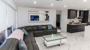A modern living room with a sectional sofa, a glass coffee table, and a kitchen area with dark cabinetry. - Modern Luxury, 2 Mi From Beach! (Fort Lauderdale)