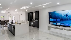 A modern kitchen with a large island, a dining area with chairs, and a large flat-screen TV mounted on the wall displaying a movie. - Modern Luxury, 2 Mi From Beach! (Fort Lauderdale)