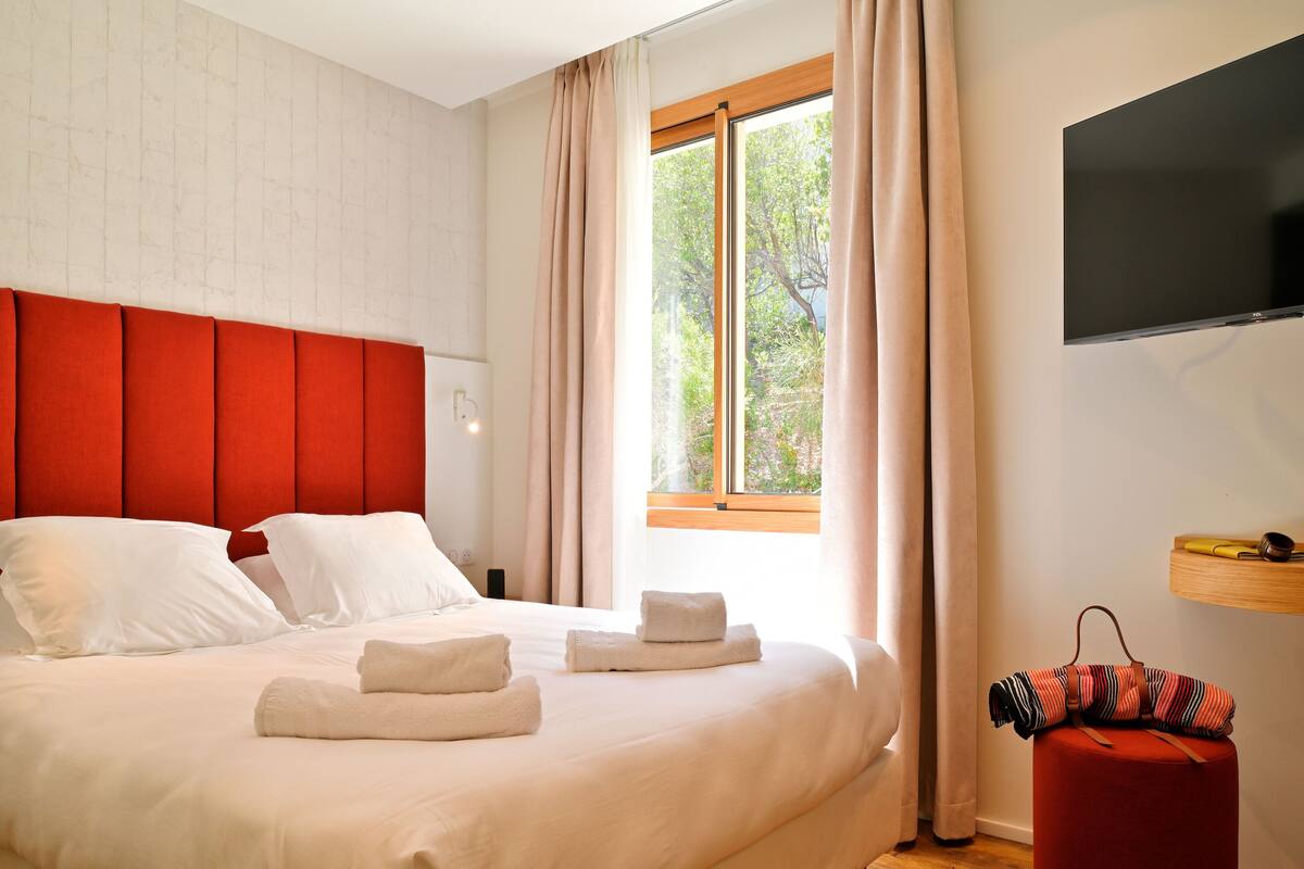 Una cama bien tendida con sábanas blancas y dos toallas, un cabecero tapizado en rojo, una ventana con cortinas y un televisor montado en la pared.