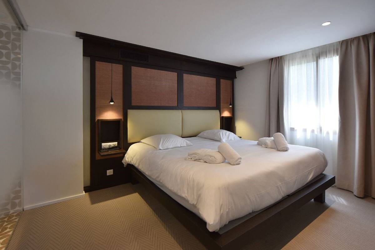 Una cama bien tendida con sábanas y toallas blancas, un cabecero con diseño de panel de madera y una mesita de noche con una lámpara.