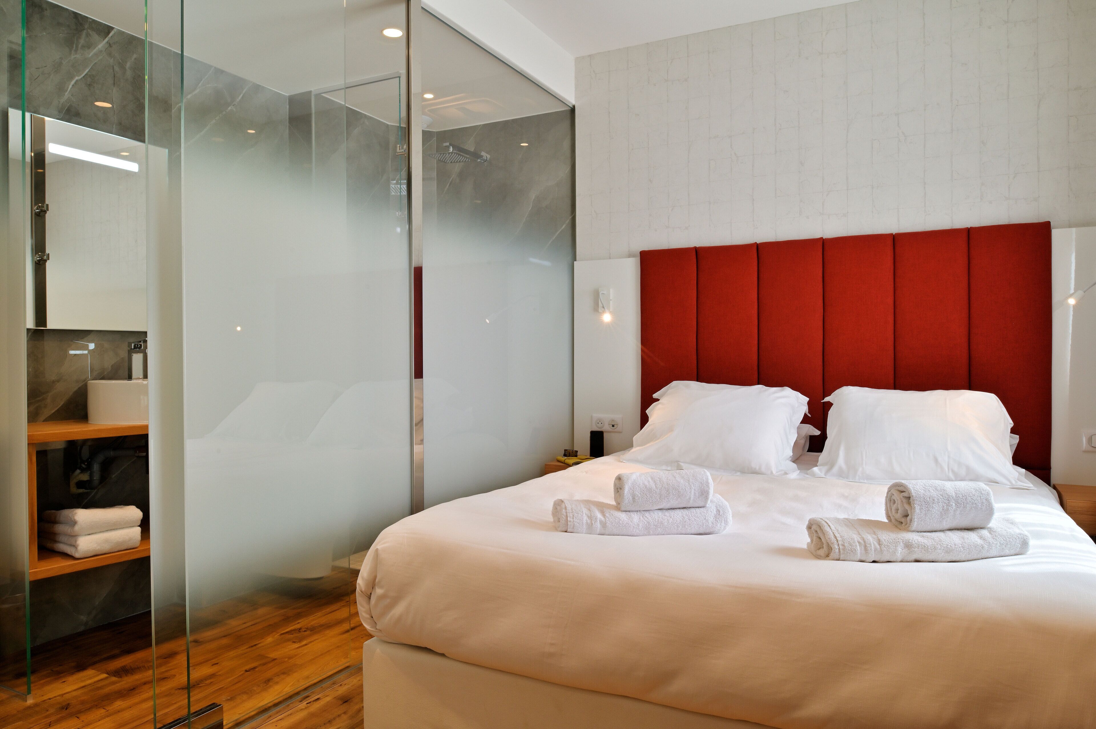 A modern hotel room with a large bed, a red headboard, and a glass partition.