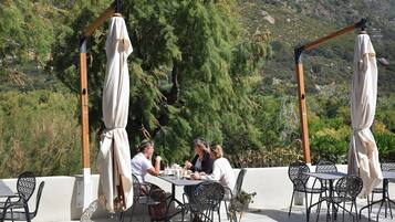 An outdoor dining area with tables and chairs, surrounded by greenery and a mountainous backdrop.