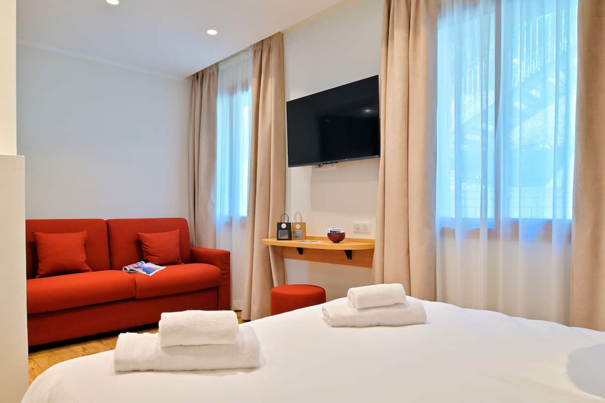 Habitación de hotel con cama, sofá rojo, televisión montada en la pared y ventana con cortinas.