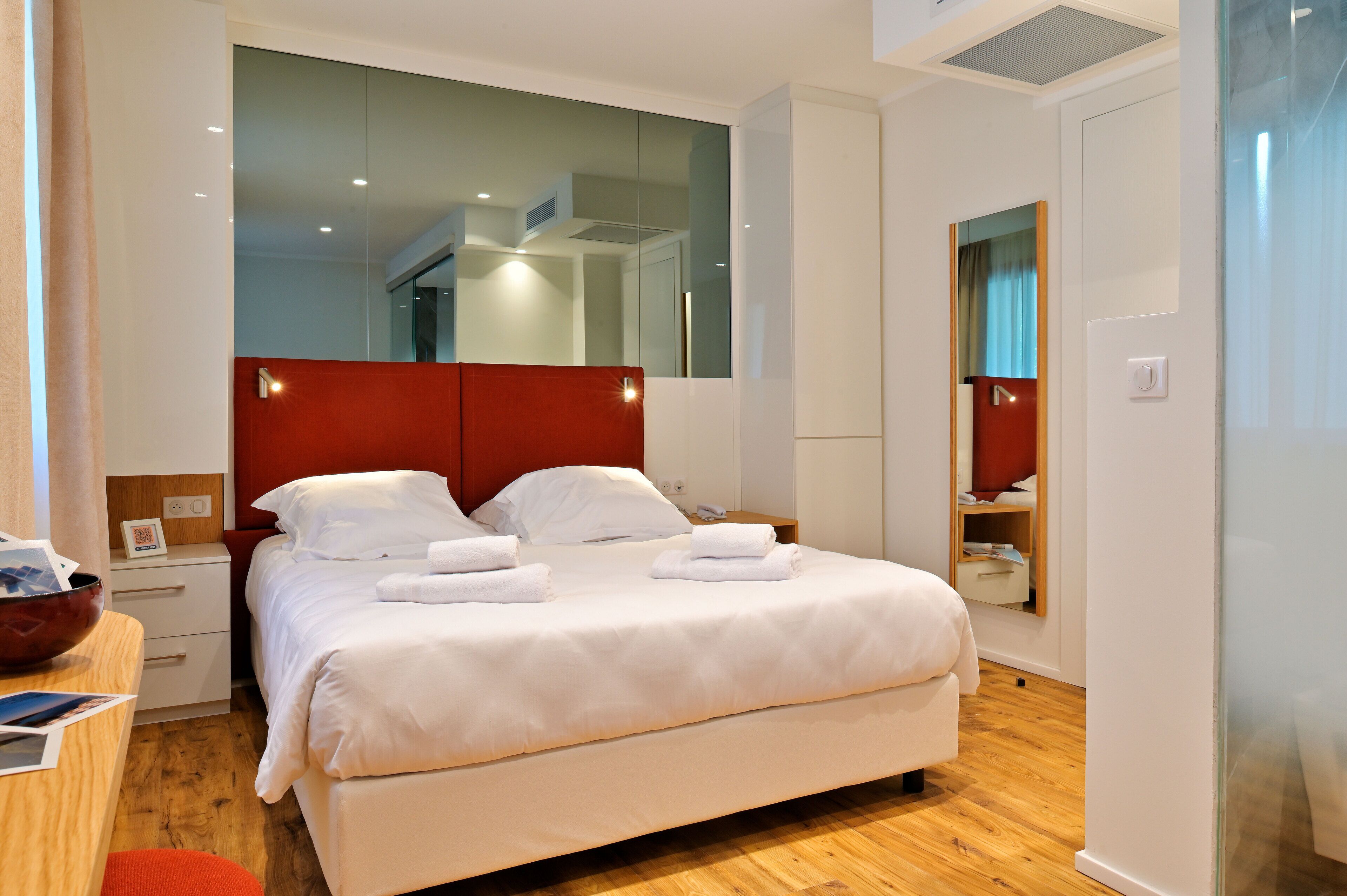 A modern hotel room with a large bed, a bedside table with a bowl, a mirror, and a wardrobe.