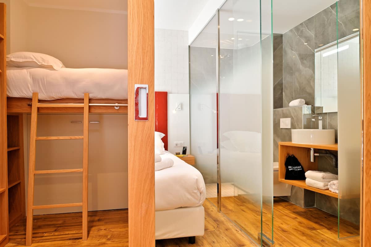 Habitación compacta con literas, una escalera de madera, una mesita de noche pequeña, un separador de vidrio y un baño con lavamanos y toallero.