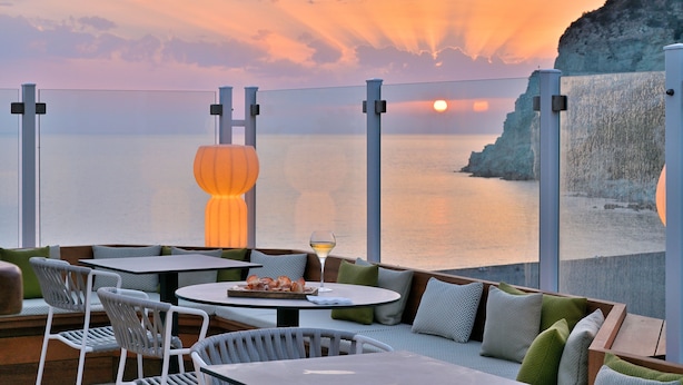A terrace with outdoor seating, a table with a glass of wine, and a view of the sunset over the sea.
