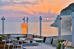 Una terraza con mobiliario exterior, una mesa con una copa de vino y vistas a la puesta de sol sobre el mar.
