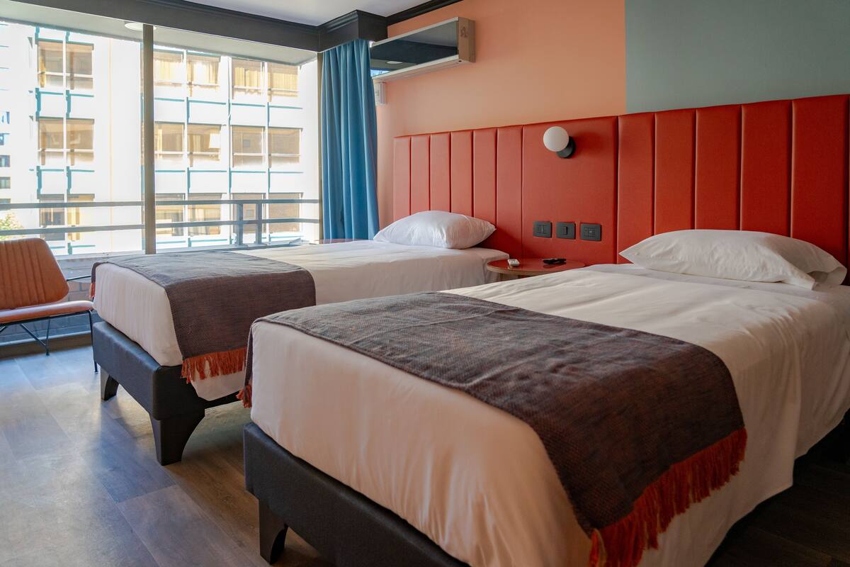 Habitación de hotel con dos camas, un ventanal con cortinas y vistas a un edificio.