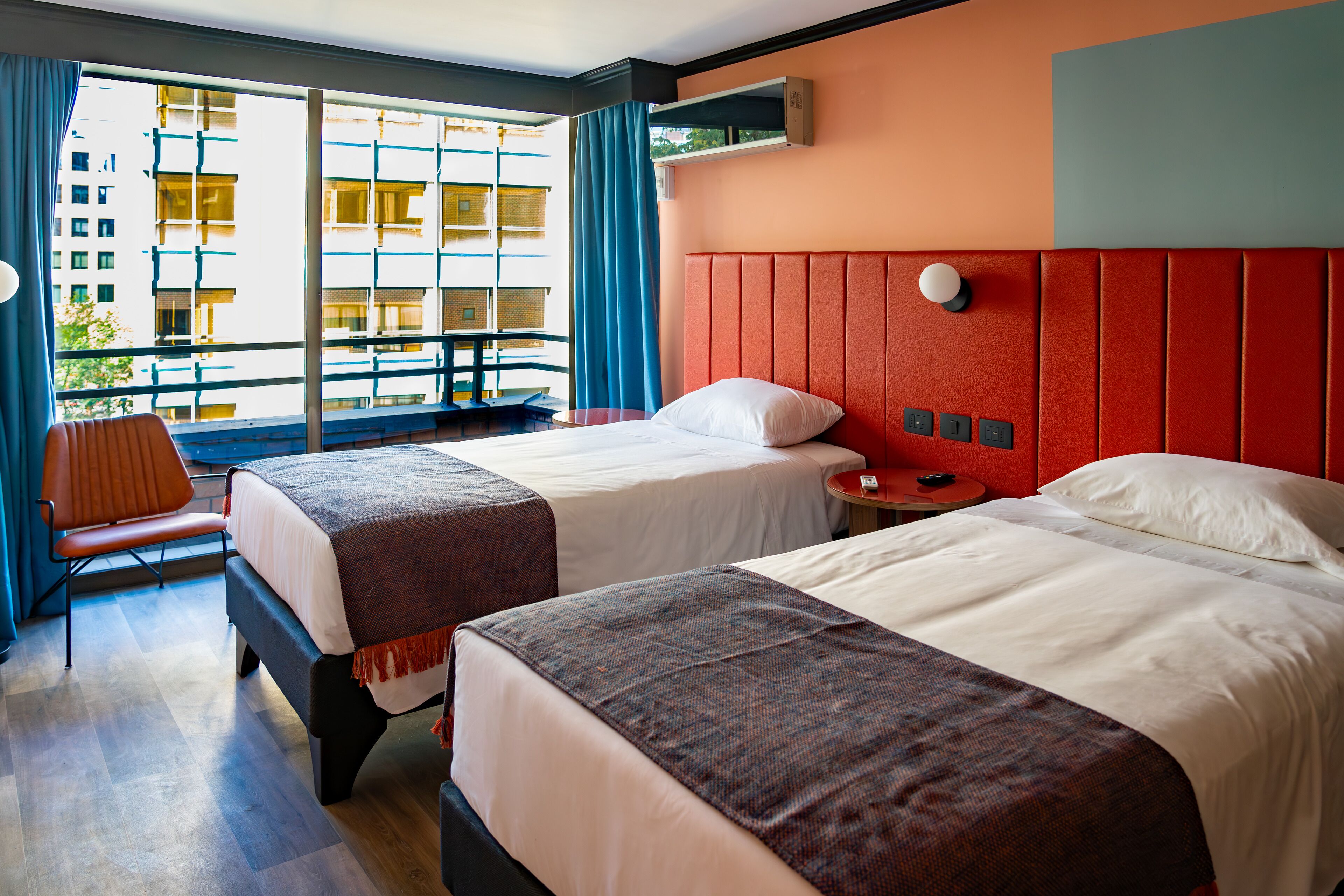A hotel room with two beds, a desk, a chair, and a large window with a view of a building.