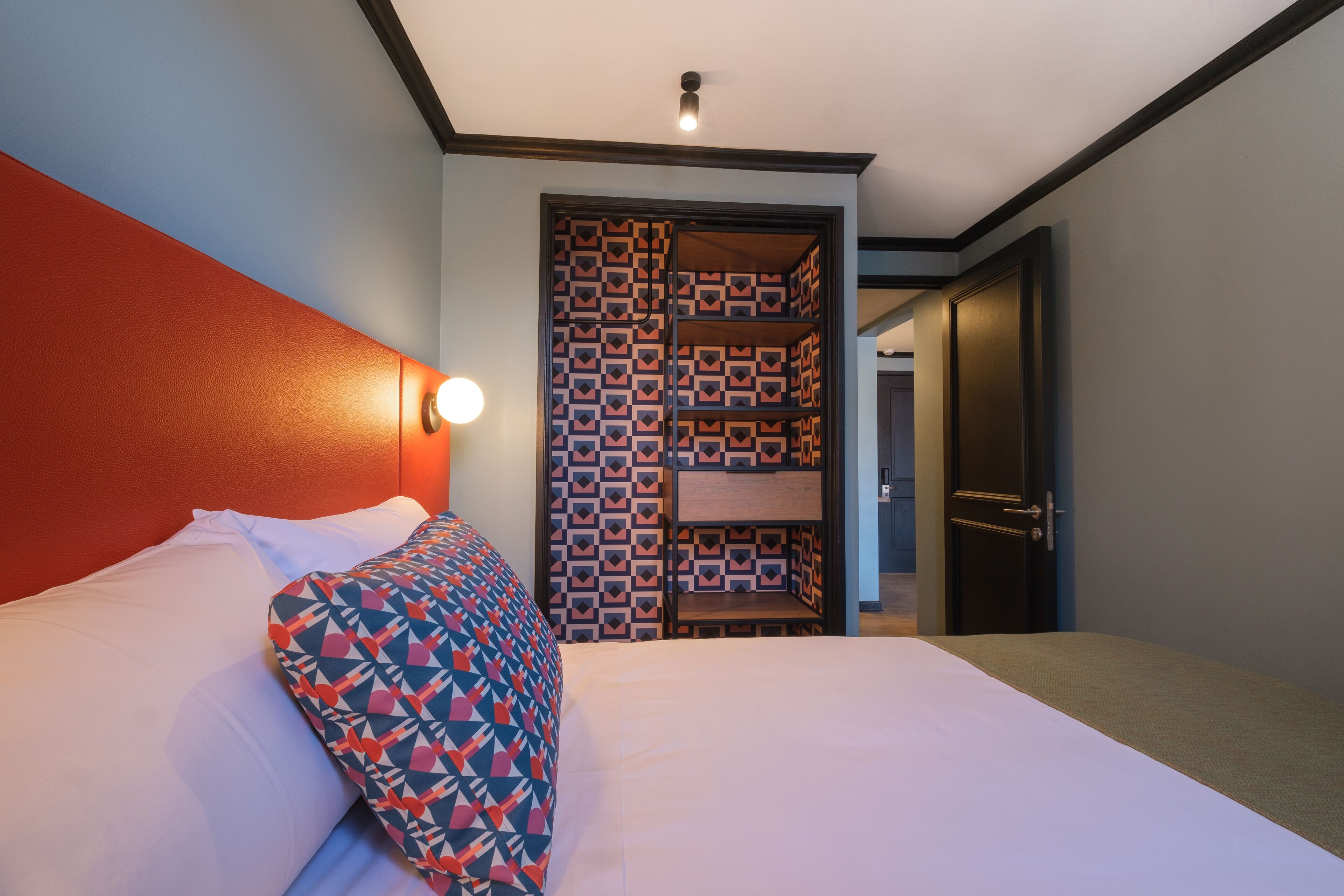 A hotel room with a bed, a headboard, a patterned wall, a door, and a light fixture.