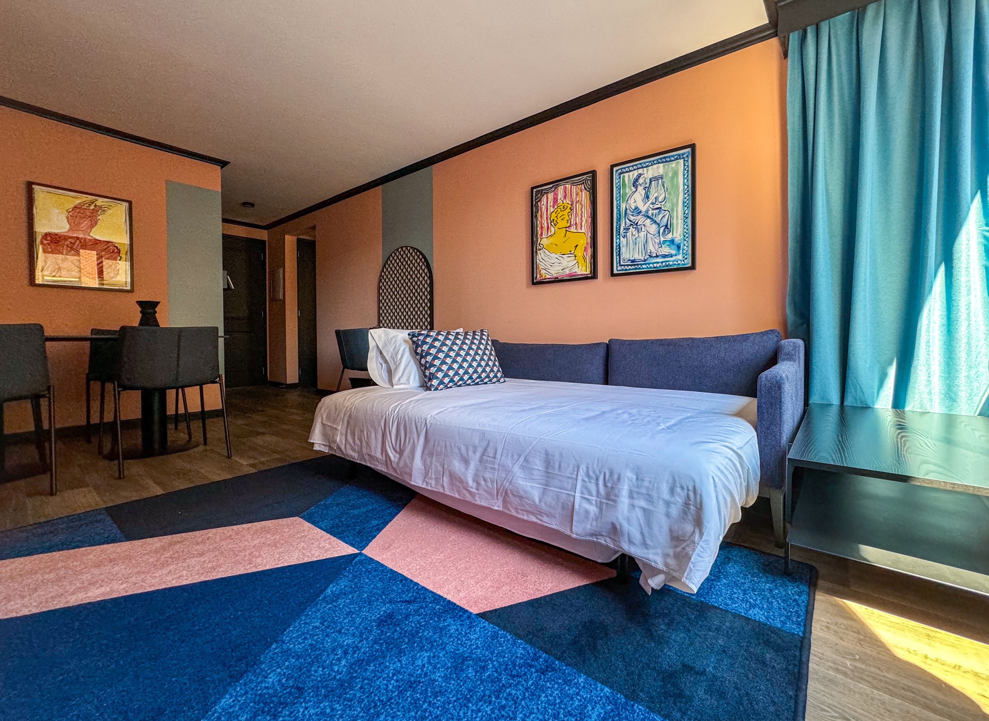 Ein modernes Hotelzimmer mit einem großen Bett, einem Schreibtisch und zwei Stühlen. An der Wand hängen zwei Bilder und es gibt ein Fenster mit Vorhängen.