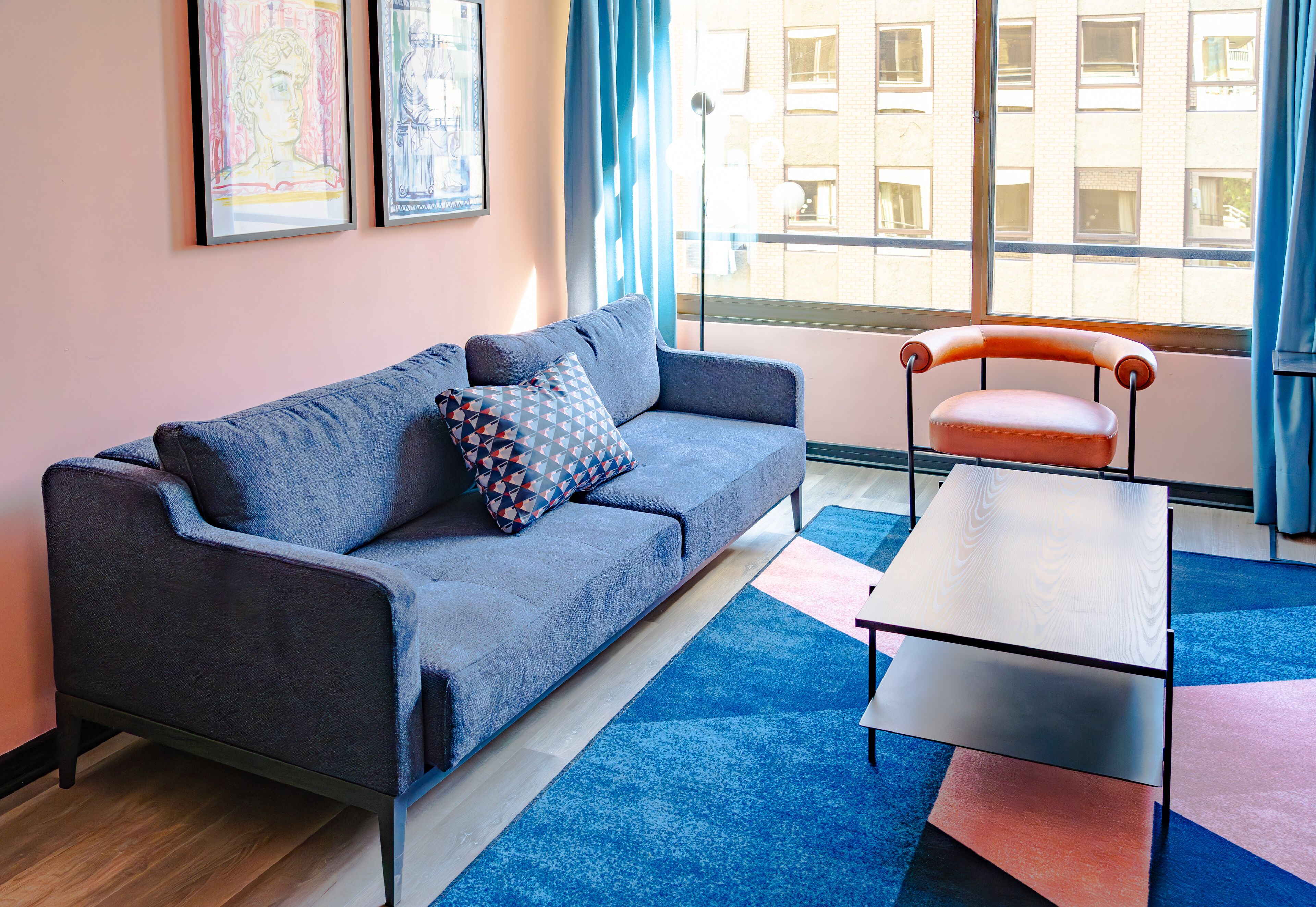 A modern living room with a grey sofa, a patterned pillow, a small table, and a view of a building through a large window.