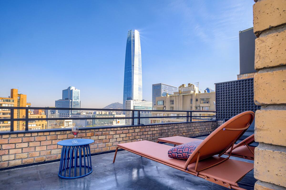 Terraza en la azotea con vistas despejadas a un rascacielos alto, un muro de ladrillo y una mesa azul con una copa de vino.