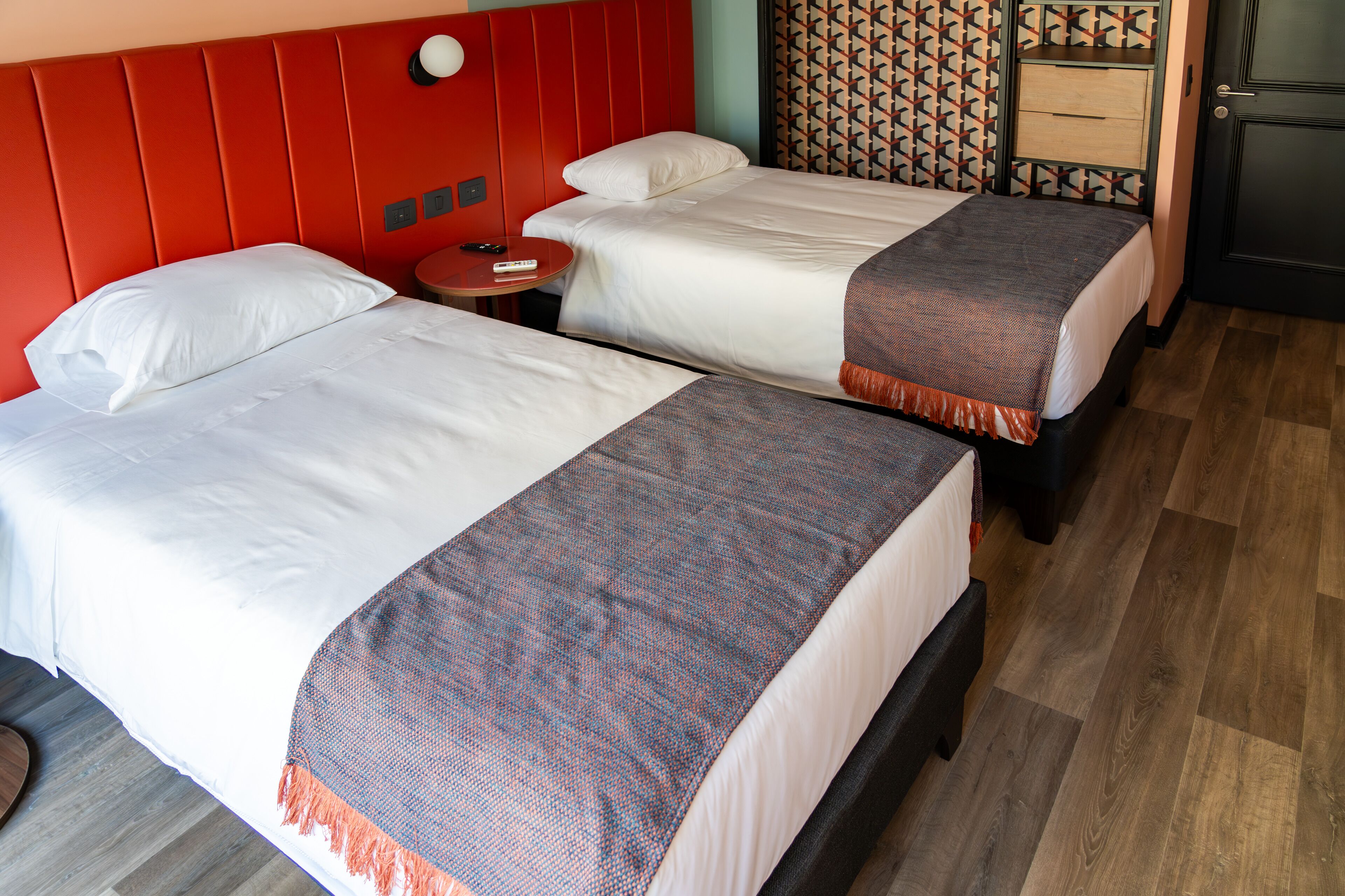 Two beds with white linens and a red headboard, a small round table with a remote control, and a patterned wall.