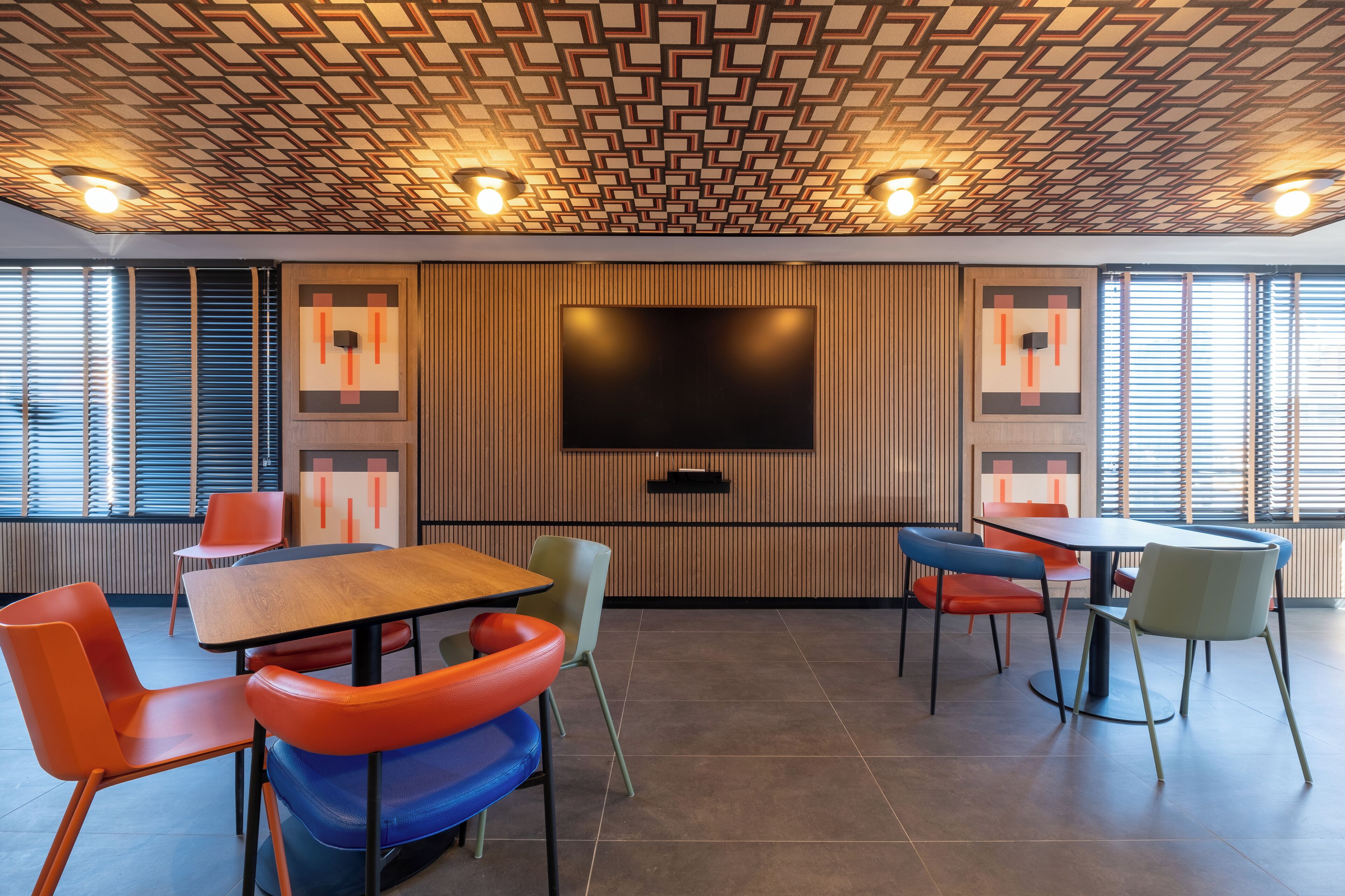 A modern conference room with a geometric patterned ceiling, a large flat-screen TV, and colorful chairs around a wooden table.