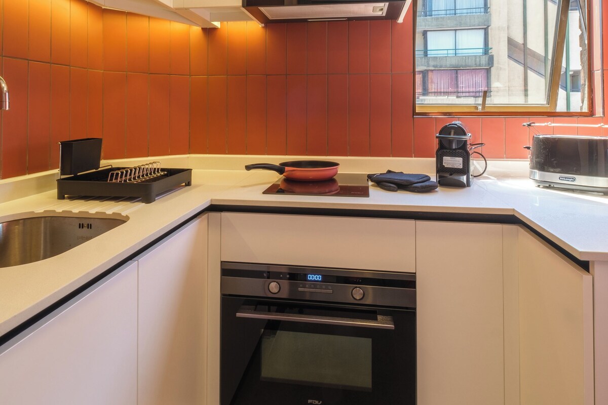 A modern kitchen with white cabinets, a built-in oven, a coffee maker, and a window with a view of buildings.