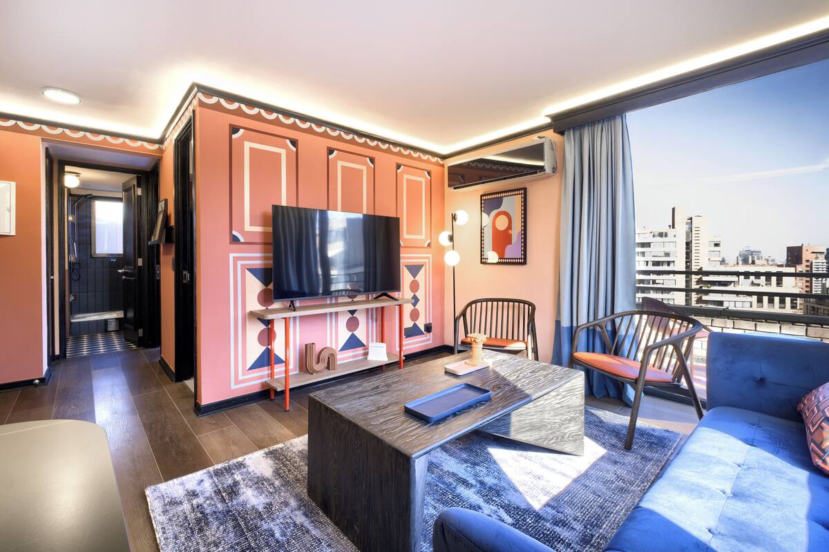 Habitación de hotel moderna con un ventanal grande, una televisión de pantalla plana, un sofá, una mesa de centro y una silla.