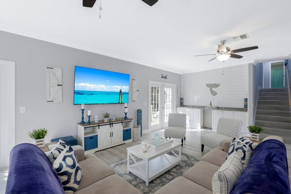 A modern living room with a large TV, a ceiling fan, and a staircase leading to the upper floor. - Barefoot Beach Private Pool & Hot Tub, Near Beach /sleeps 18 (Bonita Springs)