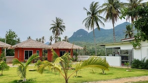 A bungalow with a thatched roof, surrounded by lush greenery and palm trees, with a mountain in the background.