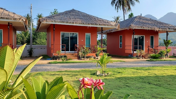 A row of red buildings with thatched roofs, surrounded by lush greenery and a clear view of mountains in the background.