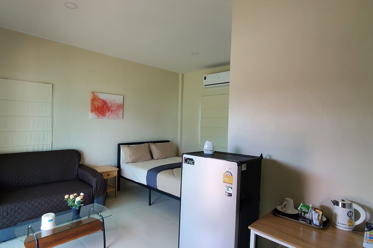 A compact room with a bed, a sofa, a small table with a vase, a refrigerator, a kettle, and a television.