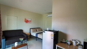 A compact room with a bed, a sofa, a small table with a vase, a refrigerator, a kettle, and a television.