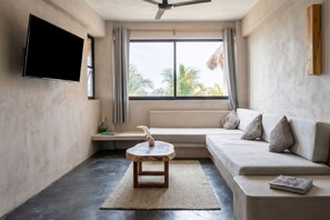 A modern living room with a flat-screen TV, a wooden coffee table, a sofa, and a large window with a view of palm trees.