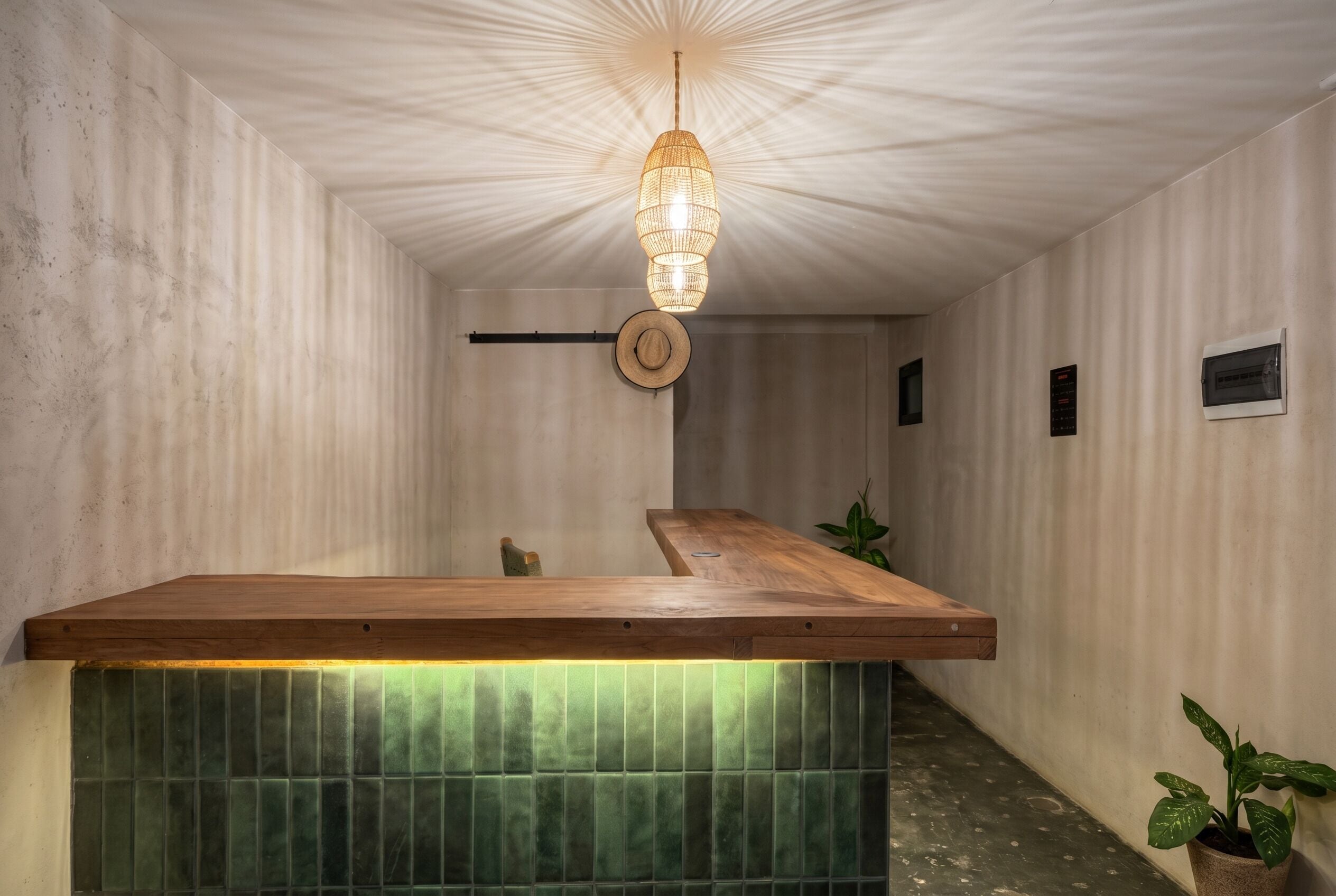 Un comptoir d’accueil en bois, avec un revêtement de carreaux verts sur le devant, un luminaire suspendu et un chapeau accroché au mur.