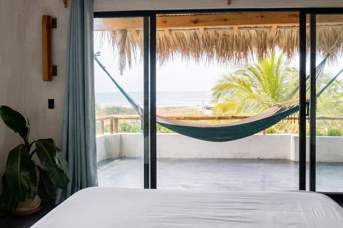 Une chambre avec un lit, une grande fenêtre, un hamac et vue sur la plage.