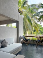 Un espacio exterior moderno con zona de estar, un sofá gris y sillones, rodeado de abundante vegetación y palmeras.