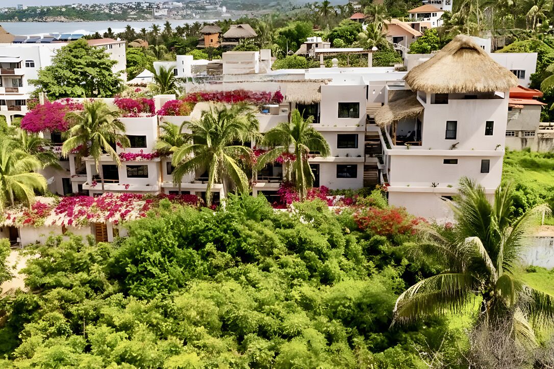 A coastal residential area with white buildings, some with thatched roofs, surrounded by lush greenery and flowering plants.