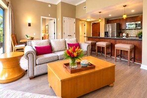A modern living room with a sofa, coffee table, and a kitchen area with bar stools and appliances. - Honua Kai | Garden View 1 BR | Free Car w/ 6+ Nights | HKH-114 (Lahaina)