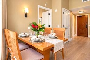 A dining area with a wooden table set for four, chairs, a vase with flowers, and a wall-mounted light fixture. - Honua Kai | Garden View 1 BR | Free Car w/ 6+ Nights | HKH-114 (Lahaina)
