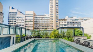 Piscina en la azotea con vistas a edificios altos, rodeada de vegetación y mobiliario exterior.