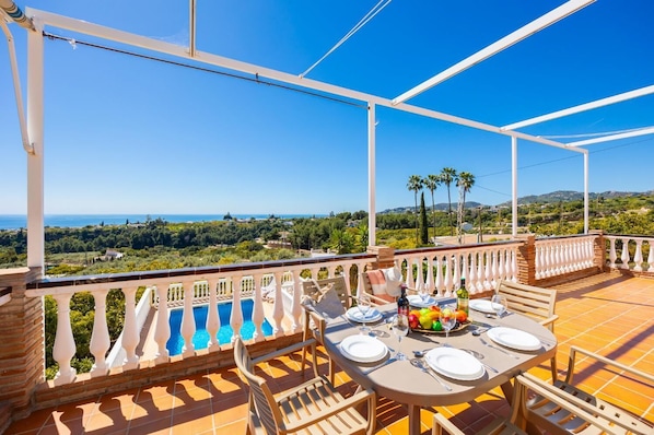 A terrace with a table set for dining, a pool, and a scenic view of the sea and hills.