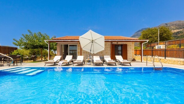 Une piscine entourée de bâtiments en pierre et d’arbres, avec des chaises longues et un parasol blanc.