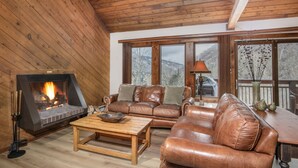 Un salon chaleureux avec une cheminée, des canapés en cuir, un plancher en bois et de grandes fenêtres offrant une vue sur les montagnes.