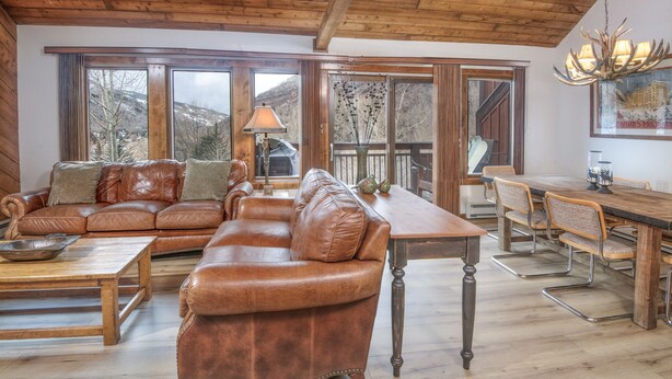 Un vaste espace de vie comprenant un canapé en cuir, une table basse en bois et une table à manger avec des chaises. De grandes fenêtres offrent une vue sur les montagnes.