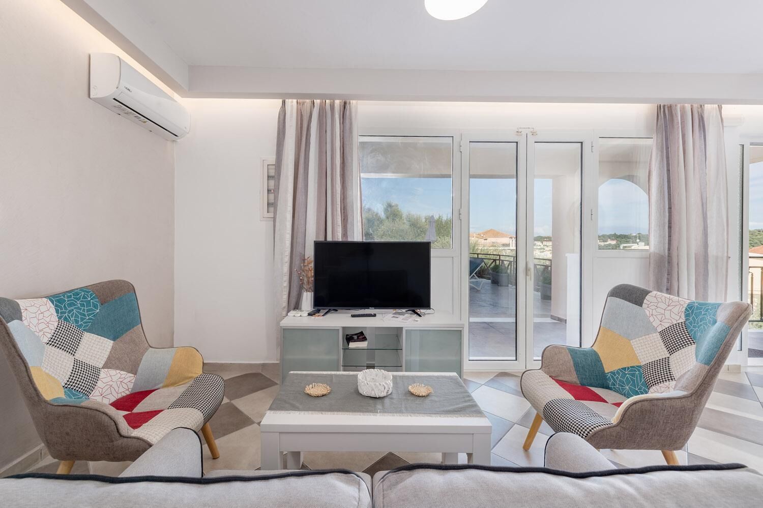 A modern living room with a flat-screen TV, two patterned armchairs, a coffee table, and a sliding glass door leading to an outdoor area.