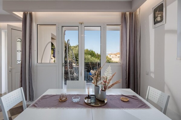 A dining area with a table set for two, a vase with dried plants, and a view of greenery outside.
